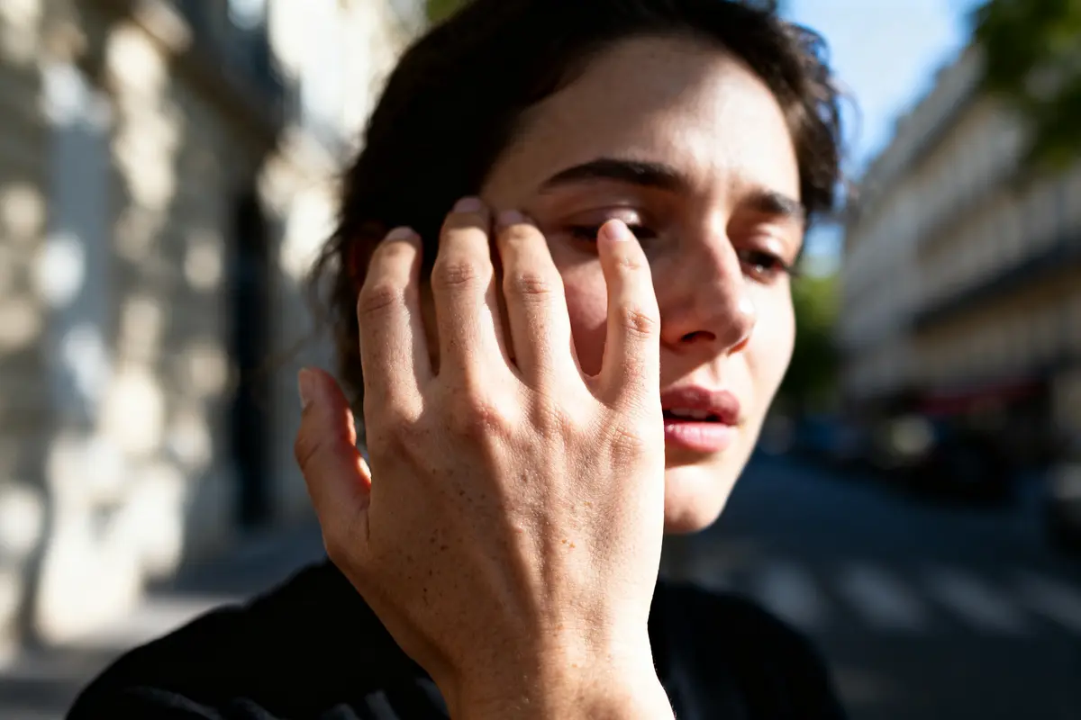 Femme touchant son visage, lumière du soleil, rue floue en arrière-plan.